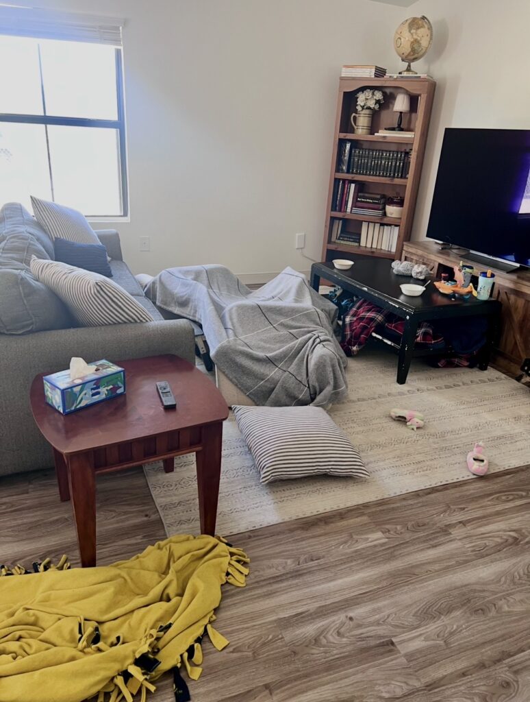 A living room fort made with blankets and pillows, showing children's playtime in a lived-in family home with everyday household items in the background.
