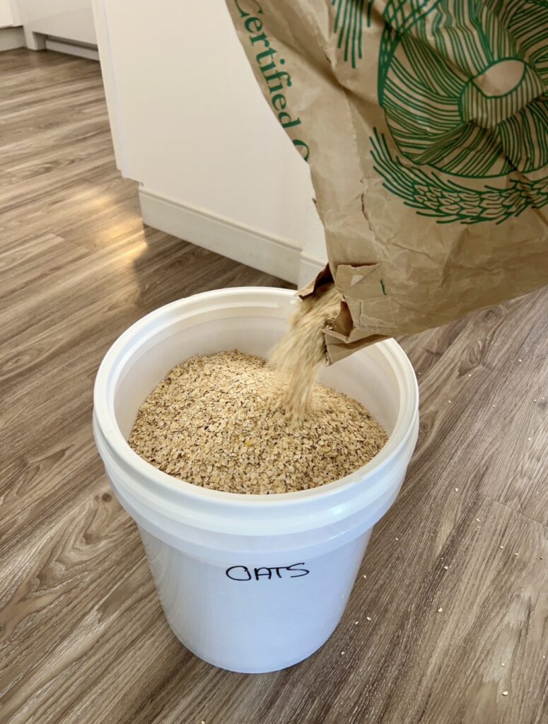 5-gallon bucket labeled oats being filled with bulk dry organic oats for emergency pantry food storage in a home kitchen.